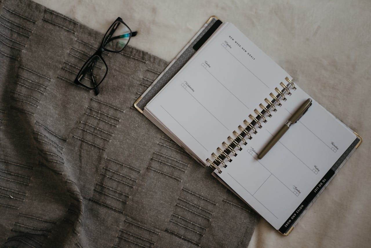about-us-02 A flatlay image of a planner, eyeglasses, and pen on a textured fabric surface.