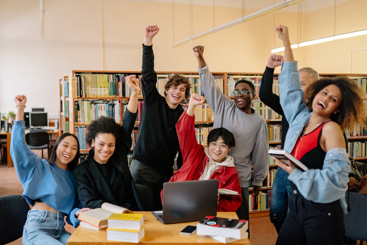 services-02 Happy diverse students in library raising hands, enjoying academic success.
